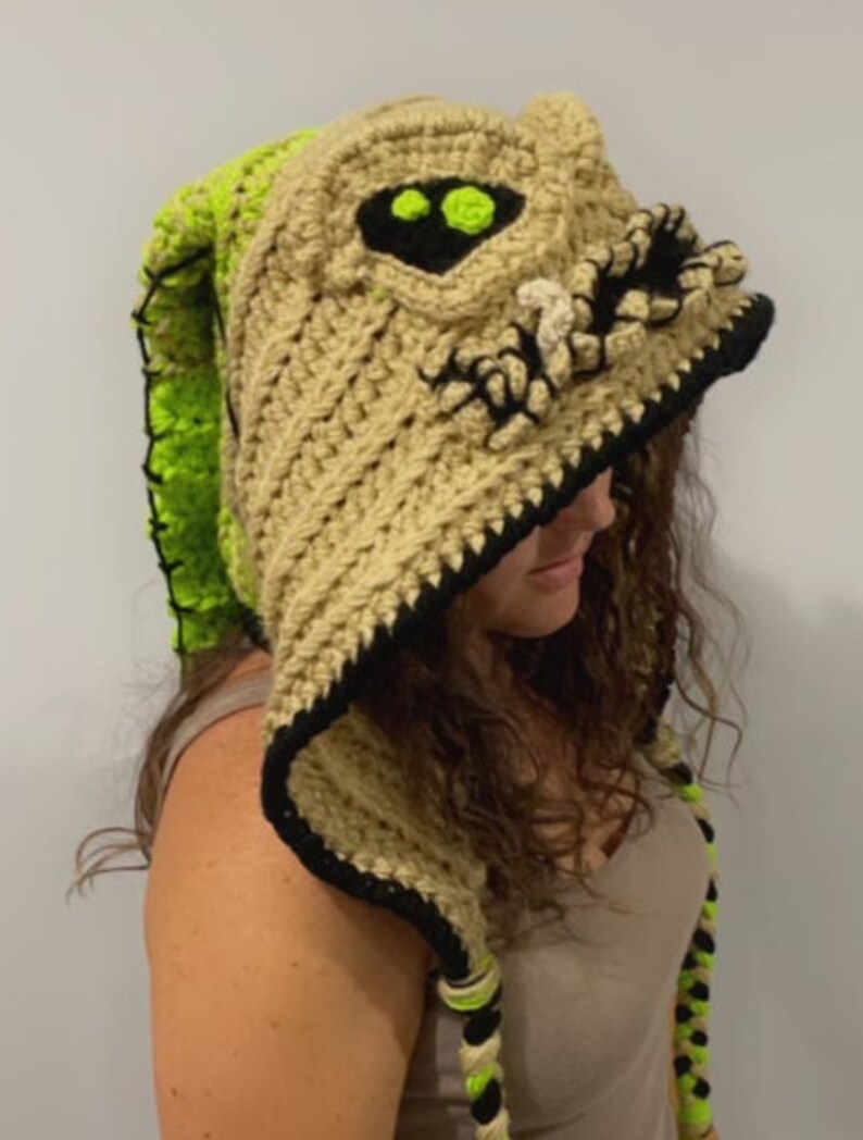 May include: A crocheted hooded hat with a beige and green color scheme. The hood features a detailed crocheted face with black eyes and a toothy grin. The hat has long, crocheted ties.