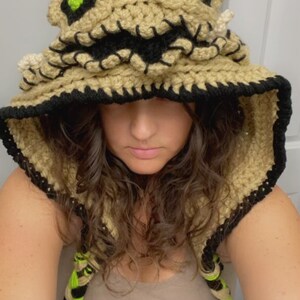 May include: A crocheted hooded hat with a brown and black color scheme. The hat features a stitched-on face with green eyes and a stitched-on mouth. The hat has a long, braided cord.