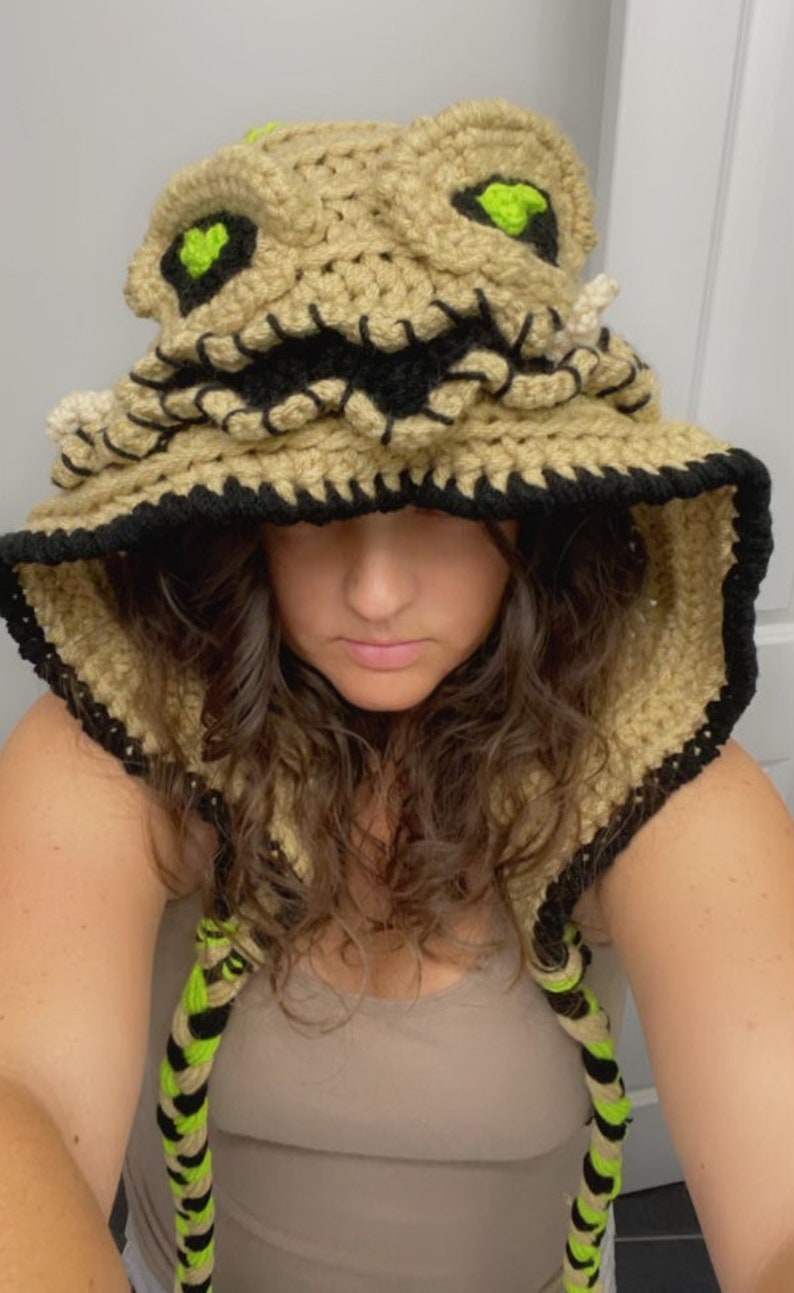 May include: A crocheted hooded hat with a brown and black color scheme. The hat features a stitched-on face with green eyes and a stitched-on mouth. The hat has a long, braided cord.