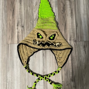 May include: A crocheted hat with a green and brown pointed top and a black and brown braided tie. The hat features a scary face with green eyes and a black mouth.
