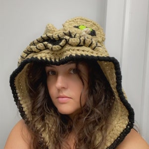 May include: A person wearing a crocheted hooded vest with a cat face design. The vest is beige with black and neon green accents.