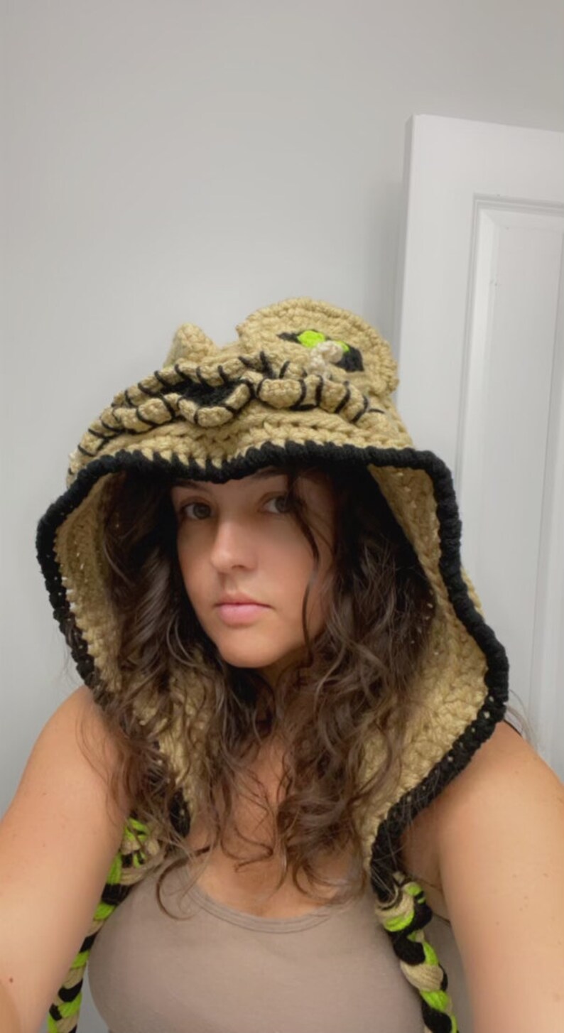 May include: A person wearing a crocheted hooded vest with a cat face design. The vest is beige with black and neon green accents.