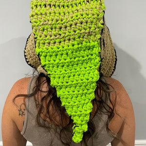 May include: A crocheted hat with a long, pointed tail. The hat is made of brown and green yarn and has a black trim. The hat is worn by a person with long, curly hair.