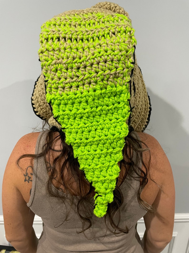 May include: A crocheted hat with a long, pointed tail. The hat is made of brown and green yarn and has a black trim. The hat is worn by a person with long, curly hair.