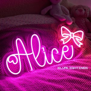 May include: A neon sign in the shape of the name "Alice" in a cursive font, with a heart detail on the "A". A neon bow is also visible. The sign is lit with a bright pink glow and rests on a pink blanket. The text "ELVALIGHTINGS" is also visible.
