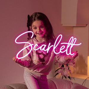 以下が含まれることがあります： 鮮やかなピンク色の筆記体で「Scarlett」と書かれたカスタムネオンサイン。ネオン管は透明な輪郭に沿ったアクリル板に取り付けられており、周囲をピンク色の光で照らす明るいディスプレイとなっています。
