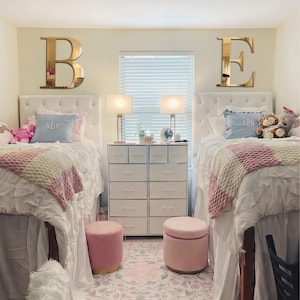 Op de afbeelding: Twee witte gecapitonneerde bedden met roze en witte gebreide dekens, lichtblauwe kussens met monogram en knuffels. Boven elk bed hangt een grote gouden spiegelletter, B en E. Tussen de bedden staat een witte ladekast met tien lades.