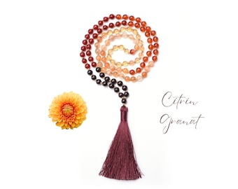 Puede incluir: Un collar de cuentas de oración o mala con un borla. Las cuentas están hechas de piedras de cornalina, citrino y granate. La borla es de color burdeos.