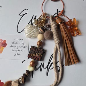 May include: A keychain featuring a tan cord, a brown faux leather tassel, and an amber-colored bear charm. It includes a white pearl-textured heart, a small brown pom-pom, and various beads, including one square bead with the text Drink Coffee Be Happy.