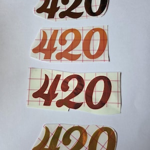 Op de afbeelding: Vier afzonderlijke stickers met het getal 420 in een gestileerd lettertype. Elke sticker wordt gepresenteerd op een wit schutvel met een rood ruitpatroon. De stickers hebben metallic afwerkingen in bruin, oranje, donkerrood en goud.