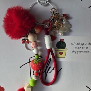 Può includere: Un portachiavi con un grande pompon rosso, un ciondolo a forma di fragola e varie perline rosse, verdi e bianche. Include un laccio rosso con avvolgimento bianco, un moschettone dorato, un ciondolo a farfalla e un cartoncino con un cactus.