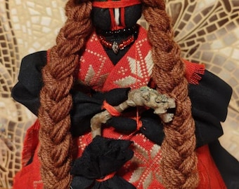 May include: A handmade doll with a red head covering and long brown braids. The doll wears a red and black patterned dress and holds a small wooden object. A black pouch is attached to the doll's clothing. The doll has a black face with red accents.