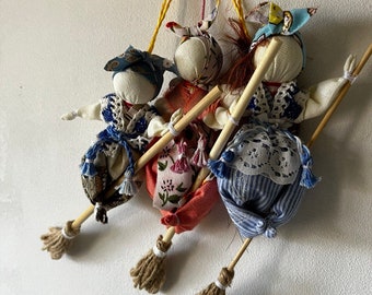 May include: Three handmade fabric dolls with colorful clothing and floral patterns are holding broomsticks. The dolls are hanging from a string and are made in a traditional style.
