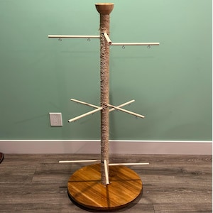 May include: A wooden display stand with a circular base and multiple horizontal arms for hanging items. The central pole is wrapped in twine, and the top has a small wooden bowl. The stand is made of light-coloured wood.