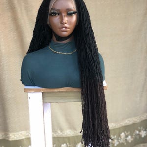 Puede incluir: Peluca negra con rastas largas y falsas, exhibida en una cabeza de maniquí. La peluca presenta una parte delantera de encaje y un efecto degradado de marrón oscuro a negro. El maniquí lleva un cuello alto verde oscuro y una cadena dorada.