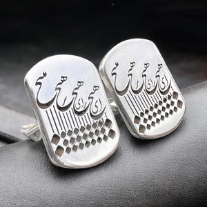 May include: A pair of silver cufflinks with an intricate Arabic calligraphy design. The cufflinks are rectangular in shape and have a checkered pattern on the back.