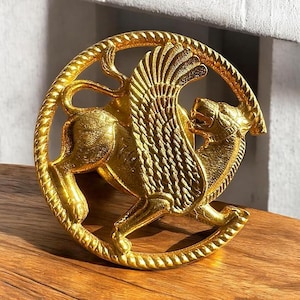 Puede incluir: Un objeto decorativo circular de color dorado con un diseño de león alado. El león está representado en una pose dinámica, con alas detalladas y una superficie texturizada. El objeto es probablemente de metal y tiene una estética antigua o vintage.
