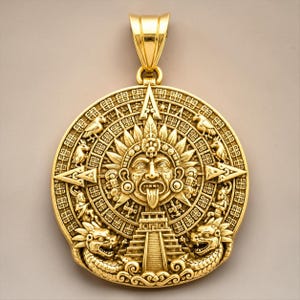 May include: A circular gold pendant featuring a detailed relief of an Aztec sun stone calendar. The design includes a central face with a protruding tongue, surrounded by intricate geometric patterns, feathered motifs, and two dragon-like figures.