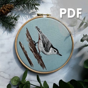 May include: Embroidered white-breasted nuthatch on a blue fabric background in a wooden embroidery hoop. The bird is perched on a brown branch.