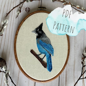 以下が含まれることがあります： 青い鳥の刺繍パターン。頭は灰色で、茶色の枝があります。刺繍は白い布に木製の輪で飾られています。テキスト「PDF PATTERN」は青い水彩の背景にあります。