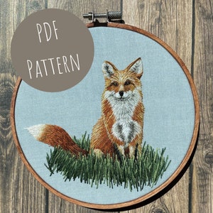May include: Embroidered fox pattern in a hoop. The fox is sitting in a patch of green grass with its tail curled behind it. The fox is orange and white with a black nose and eyes.