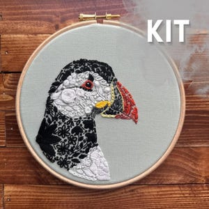 May include: An embroidery kit featuring a puffin portrait stitched onto light grey fabric within a circular wooden hoop. The bird is composed of intricate black, white, and red floral patterns. The word KIT is printed in white in the upper right.