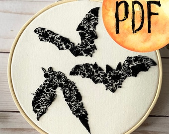 May include: A white embroidery hoop with three black bats made of floral embroidery. The bats are arranged in a cluster with their wings spread out. The hoop is on a wooden surface.