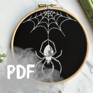 May include: White embroidered spider hanging from a web on a black fabric background in a wooden embroidery hoop. The spider is in a misty, cloudy design.