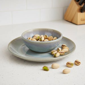 May include: A round ceramic bowl and matching plate set featuring a smooth, glossy glaze in shades of muted blue and grey. The bowl is filled with light brown pistachio nuts, and several additional pistachios are scattered on the plate surface.
