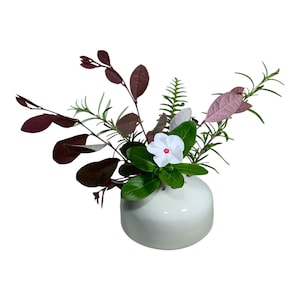 May include: A white ceramic bud vase with a rounded base and multiple openings, containing an arrangement of green fern fronds, rosemary sprigs, dark purple foliage, and a single white flower with a small red center.