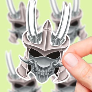 Könnte beinhalten: Ein Aufkleber mit einer detaillierten Illustration eines Samurai-Helms mit schädelähnlichem Gesicht. Der Helm ist hauptsächlich silber und grau, mit scharfen, gebogenen Hörnern und einem bedrohlichen Ausdruck. Der Aufkleber wird von einer Hand vor einem hellgrünen Hintergrund gehalten.