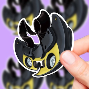 以下が含まれることがあります： 黒と黄色の漫画風デザインの、型抜きステッカー。ステッカーは白い縁取りがあり、同様のステッカーのぼやけた背景に対して手で持たれています。デザインはユニークで機械的な美しさを持っています。