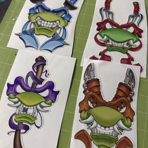 May include: Four colorful die-cut stickers featuring stylized turtle-like faces with weapons. Each sticker has a different color scheme: blue, red, purple, and brown. The stickers are on white backing paper, placed on a green cutting mat.