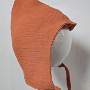 May include: A rust-colored bonnet made of a textured, crinkled fabric. The bonnet features a pointed top, a curved brim that frames the face, and two long, matching fabric ties extending from the bottom to secure the head covering in place.