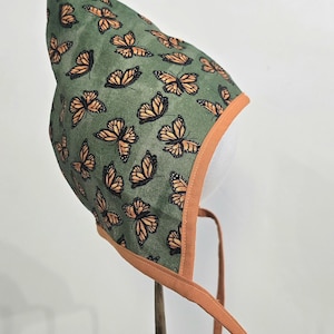 May include: A pointed bonnet featuring a green fabric base with a repeating pattern of orange and black monarch butterflies. The bonnet is finished with a solid orange fabric trim along the front edge and includes two long orange fabric ties.