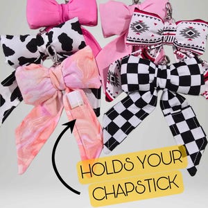 May include: A collection of bow-shaped chapstick holders in various patterns and colors. Designs include pink, cow print, marble, geometric, and checkered. Each bow has a metal clip and the text "HOLDS YOUR CHAPSTICK".