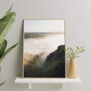May include: A framed photograph of a mountain valley with fog rolling through it. The valley is green and the fog is white. The photo is in a light wood frame.