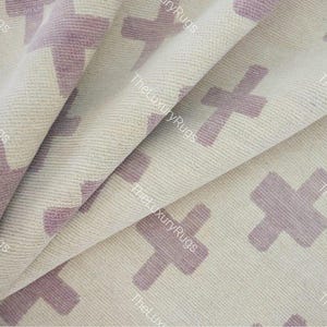 Puede incluir: Primer plano de una alfombra color crema con un patrón repetido de cruces lavanda. La alfombra está doblada, mostrando la textura y el diseño. The Luxury Rugs está impreso en la alfombra.