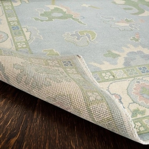May include: A light blue area rug with a floral pattern in green, pink, and cream. The rug's edge is folded over, revealing the underside's woven texture. The rug is placed on a dark brown wooden surface.