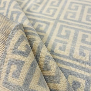 May include: A rolled-up area rug with a repeating geometric pattern in light blue and beige. The design features a Greek key motif, creating a textured surface. The rug's edges are visible, showcasing the layered construction.