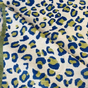 Peut inclure: Un textile plié avec un fond crème, orné de motifs de taches de léopard bleues et vert olive. Le bord est bordé d'une bordure vert olive uni. Le texte "Design A" est dans un rectangle arrondi vert et blanc.