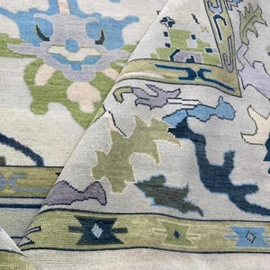 Könnte beinhalten: Gefalteter, hellfarbiger Teppich mit detailliertem Muster. Der Teppich zeigt ein zentrales Design mit blauen und grünen blütenartigen Formen. Der Rand hat ein geometrisches Muster in Grün-, Blau- und Beigetönen. Der Teppich besteht aus weichem Material.