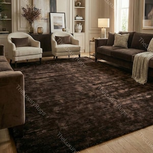 May include: A large, dark brown area rug with a textured, slightly shimmering surface. The rug is the focal point of a living room, complementing the neutral-toned furniture and decor. The room features two armchairs, a sofa, and wooden flooring.