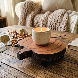 May include: A lit candle in a cream-colored ceramic container sits on a wooden, pumpkin-shaped tray. The tray is on a rustic, wooden table, with a book and compass nearby. Decorative sprigs and leaves add to the fall theme.