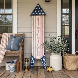May include: A wooden rocket-shaped decoration with a patriotic theme. The top is blue with white stars, the body has red and white stripes, and the number "1776" is displayed. It's part of a porch decor setup with a bench, pillows, and a lantern.
