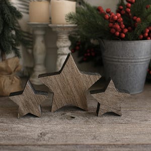 May include: Three wooden star decorations with a rustic wood grain finish. The stars are different sizes and arranged on a wooden surface. The background includes candles, greenery, and a metal bucket, suggesting a holiday theme.