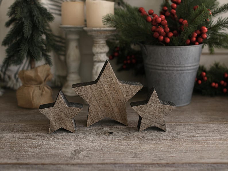 May include: Three wooden star decorations with a rustic wood grain finish. The stars are different sizes and arranged on a wooden surface. The background includes candles, greenery, and a metal bucket, suggesting a holiday theme.