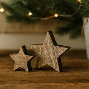 May include: Two rustic wooden star decorations, one large and one small, with a natural wood grain finish. The stars are placed on a wooden surface, with a blurred background of greenery and fairy lights, suggesting a holiday theme.