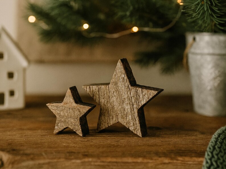 May include: Two rustic wooden star decorations, one large and one small, with a natural wood grain finish. The stars are placed on a wooden surface, with a blurred background of greenery and fairy lights, suggesting a holiday theme.