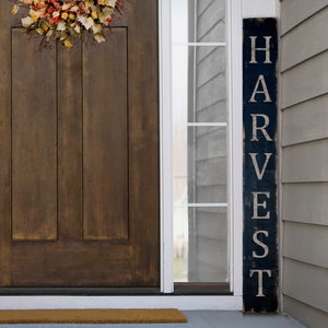 May include: A dark blue wooden sign with the word "HARVEST" in white lettering stands next to a brown wooden door. A fall wreath with orange, yellow, and brown leaves hangs above the door. The door has a silver handle and lock.